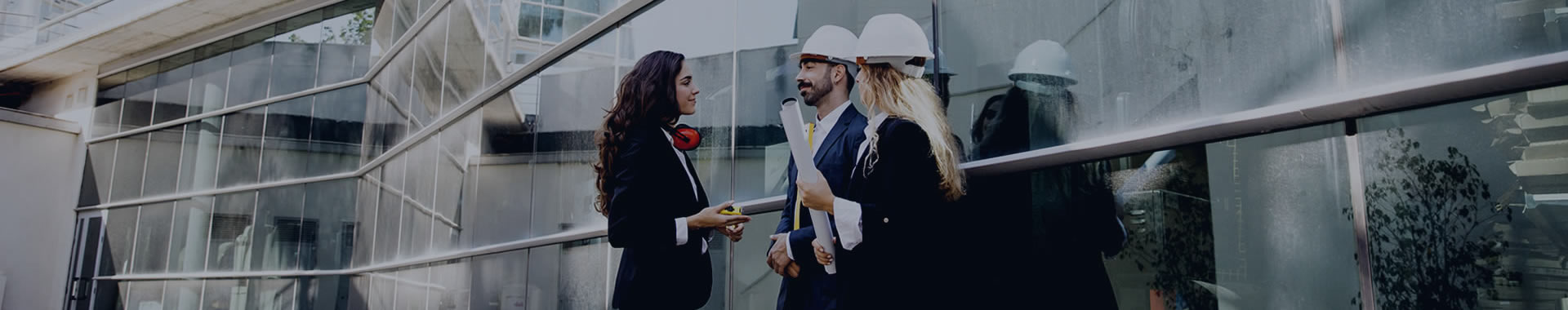 A man and woman wearing construction hats talking to a business woman outside of a skyrise that's under construction.